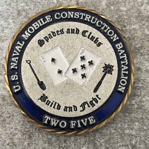 May include: A circular challenge coin with a gold-toned rope edge, a blue enamel border, and a silver-toned center. It features the text U.S. NAVAL MOBILE CONSTRUCTION BATTALION, Spades and Clubs, Build and Fight, and TWO FIVE, with icons of playing cards, a shovel, and a mace.