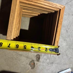 May include: A square wooden frame with a light brown natural wood grain finish. A yellow measuring tape is positioned across the top edge, showing a width measurement of approximately 8 inches from the outer edge to the inner corner.