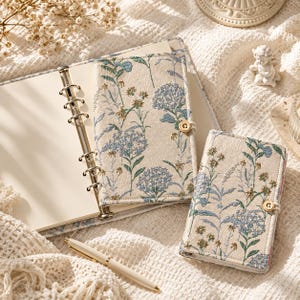 May include: A cream-colored fabric planner with a floral pattern in shades of blue and green, featuring a six-ring metal binder mechanism, a button closure, and a matching smaller notebook, accompanied by a white pen on a textured fabric surface.