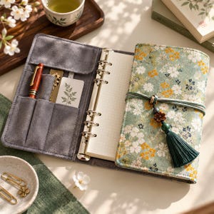 May include: A six-ring binder planner with a sage green floral fabric cover, featuring a dark green tassel and cord closure. The interior is lined with gray suede-like material, including pen slots, card pockets, and white grid-lined paper pages.