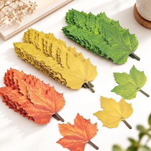 May include: A collection of artificial maple leaf decorations in three distinct color groups: vibrant green, golden yellow, and autumn orange. Each leaf features a realistic serrated edge, detailed vein patterns, and a small brown stem at the base.