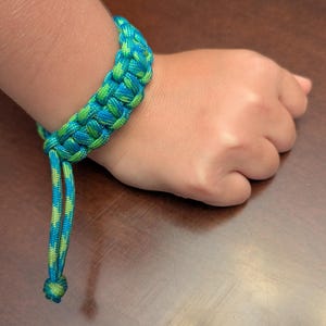 May include: A paracord bracelet featuring a woven pattern in vibrant shades of blue and lime green. The bracelet is constructed from a single continuous cord, with two loose ends hanging down, each finished with a small knot at the very tip.