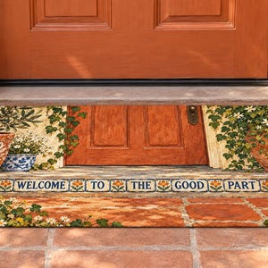 May include: A rectangular doormat featuring a printed scene of a terracotta-colored door surrounded by climbing ivy and potted plants. The text "WELCOME TO THE GOOD PART" is centered on a decorative border with small orange flower illustrations.