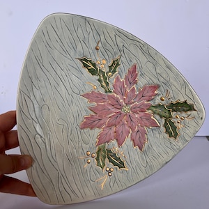 May include: A triangular ceramic plate with a textured, light gray wood-grain finish. The center features a hand-painted pink poinsettia flower with dark green holly leaves and small gold-colored berries, accented with delicate gold outlines.