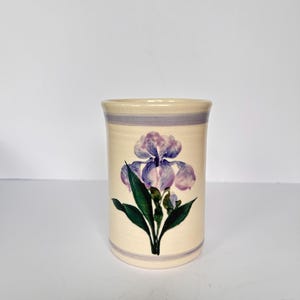 May include: A cylindrical ceramic vase with a cream-colored base featuring a hand-painted purple iris flower with green leaves. The vase has horizontal purple bands encircling the top and bottom rims, providing a simple and clean aesthetic.