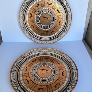 May include: Two round decorative plates featuring a metallic gold finish with black illustrations of leaping gazelles and stylized trees. The borders are decorated with a black and white Greek key pattern and concentric geometric bands.