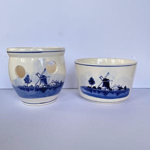 Puede incluir: Una maceta de cerámica con dos aberturas circulares y un cuenco a juego, ambos con base blanca y un diseño de paisaje pintado en azul que muestra un molino de viento, árboles y casas junto a la orilla, con bordes decorados en azul.
