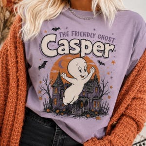 May include: A light purple t-shirt featuring a graphic of the cartoon character Casper the Friendly Ghost floating in front of a haunted house. The design includes an orange moon, black bats, stars, and the text THE FRIENDLY GHOST Casper.