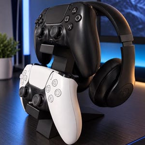 May include: A black plastic gaming stand holding one black wireless controller, one white wireless controller, and a pair of black over-ear headphones. The stand features tiered mounts for the controllers and an arched top for the headphones.