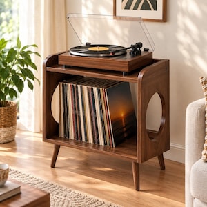 May include: A dark wood record player stand with four tapered legs and circular side cutouts. The top surface holds a black turntable with a clear dust cover, while the lower shelf contains a collection of vinyl records in sleeves.