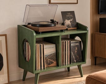 Puede incluir: Un mueble para tocadiscos de madera verde oscuro con cuatro patas cónicas y dos aberturas laterales circulares. La superficie superior sostiene un tocadiscos de madera con tapa transparente. Dos estantes interiores contienen discos de vinilo.
