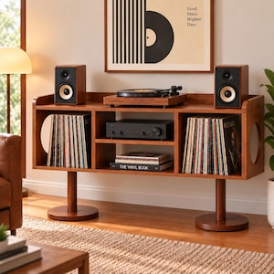 May include: A wooden media console with two circular side cutouts, supported by two cylindrical pedestals. It holds a turntable, two speakers, an amplifier, and vinyl records. A book titled The Vinyl Book is on the middle shelf.