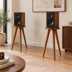 May include: Two wooden speaker stands, each featuring a circular top and three tapered legs, supporting rectangular bookshelf speakers with black front panels. The stands are crafted from a medium-toned wood, matching the finish of the speaker cabinets.