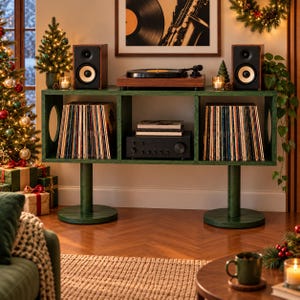 May include: A dark green wooden media console with three open storage compartments, supported by two cylindrical pedestals. The unit holds two rows of vinyl records and a black audio amplifier, with a turntable and speakers on top.
