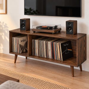 May include: A dark wood media console with four tapered legs, featuring two open storage compartments filled with vinyl records. A turntable sits on top, flanked by two matching dark wood speakers, positioned below a wall-mounted television screen.
