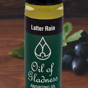 May include: A clear glass bottle filled with light yellow liquid, featuring a black cap and a dark green label. The label displays the text Latter Rain, Oil of Gladness, ANOINTING OIL, and 1/2 oz. Bottle, along with a white droplet icon.