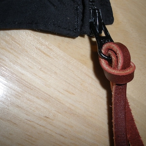 May include: A black zipper pull featuring a dark brown leather cord knotted through the metal slider. The zipper is attached to a black fabric edge, showcasing the contrast between the dark textile and the natural leather texture.