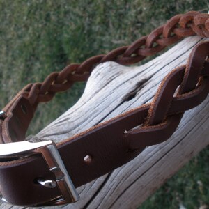 Puede incluir: Un collar de cuero marr&oacute;n oscuro con un dise&ntilde;o trenzado a lo largo de la correa. El collar incluye una hebilla de metal plateado pulido y una anilla en D a juego para la sujeci&oacute;n, mostrando un acabado liso en la correa y bordes texturizados.