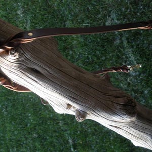 Puede incluir: Una correa de cuero marr&oacute;n oscuro con una secci&oacute;n trenzada cerca del asa y un mosquet&oacute;n giratorio de metal en el extremo. La correa est&aacute; fabricada con una tira de cuero plana, un remache de color lat&oacute;n y una anilla met&aacute;lica circular.