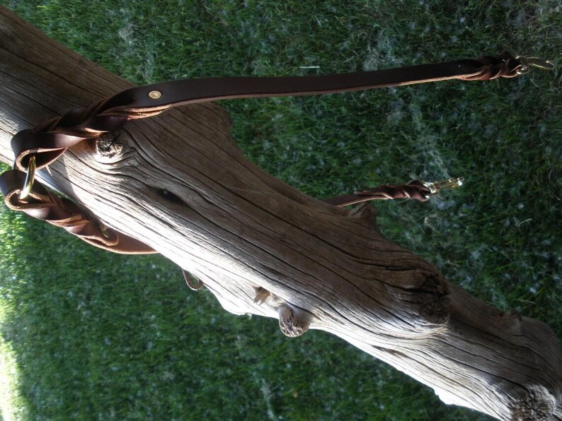 Puede incluir: Una correa de cuero marr&oacute;n oscuro con una secci&oacute;n trenzada cerca del asa y un mosquet&oacute;n giratorio de metal en el extremo. La correa est&aacute; fabricada con una tira de cuero plana, un remache de color lat&oacute;n y una anilla met&aacute;lica circular.