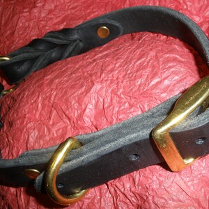 Puede incluir: Un collar de cuero negro con una secci&oacute;n trenzada, una hebilla de metal dorado, una anilla en D a juego y remaches dorados. El collar est&aacute; fabricado con tiras de cuero negro mate grueso aseguradas con herrajes met&aacute;licos.