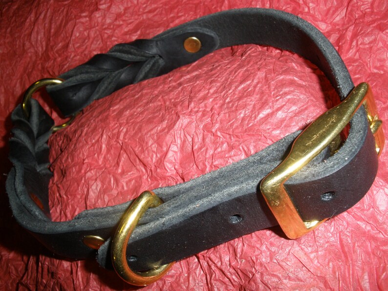 Puede incluir: Un collar de cuero negro con una secci&oacute;n trenzada, una hebilla de metal dorado, una anilla en D a juego y remaches dorados. El collar est&aacute; fabricado con tiras de cuero negro mate grueso aseguradas con herrajes met&aacute;licos.