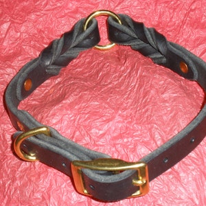 Puede incluir: Un collar de cuero negro con una secci&oacute;n trenzada, herrajes de metal dorado y cierre de hebilla. El dise&ntilde;o incluye un anillo circular en la parte superior y remaches de color cobre que aseguran las correas de cuero en forma circular.