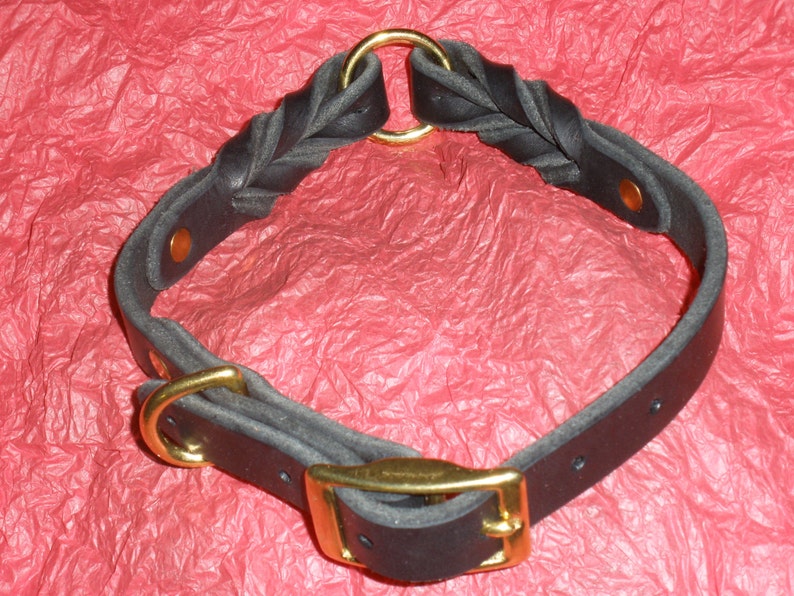 Puede incluir: Un collar de cuero negro con una secci&oacute;n trenzada, herrajes de metal dorado y cierre de hebilla. El dise&ntilde;o incluye un anillo circular en la parte superior y remaches de color cobre que aseguran las correas de cuero en forma circular.