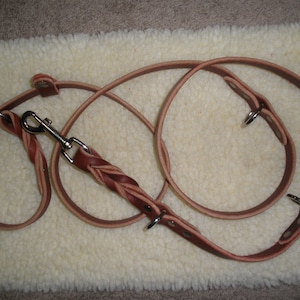 Puede incluir: Una correa de perro de cuero marrón con una sección trenzada y un clip de metal. La correa está enrollada sobre una superficie blanca y esponjosa.