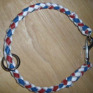 Puede incluir: Un collar trenzado para mascotas con un patr&oacute;n repetitivo de tiras de cuero en colores rojo, blanco y azul. El collar incluye una argolla met&aacute;lica circular en un extremo y un mosquet&oacute;n giratorio de metal plateado en el otro extremo.