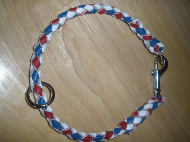 Puede incluir: Un collar trenzado para mascotas con un patr&oacute;n repetitivo de tiras de cuero en colores rojo, blanco y azul. El collar incluye una argolla met&aacute;lica circular en un extremo y un mosquet&oacute;n giratorio de metal plateado en el otro extremo.