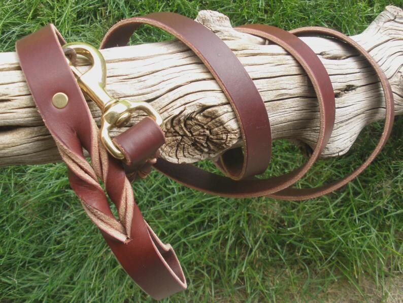Pu&ograve; includere: Un guinzaglio in pelle marrone scuro con una sezione dell'impugnatura intrecciata e un moschettone girevole in ottone lucido. La cinghia &egrave; realizzata in un unico pezzo di pelle con un rivetto metallico che fissa l'anello vicino all'ottone.
