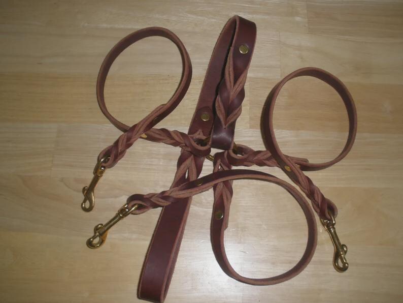 Pu&ograve; includere: Un guinzaglio multiplo in pelle marrone con un design centrale intrecciato e tre guinzagli singoli fissati a moschettoni girevoli in ottone. Il sistema &egrave; fissato con rivetti metallici e include un'impugnatura all'estremit&agrave; superiore.