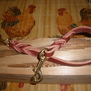 May include: A braided, burgundy leather dog leash with brass-colored metal clasps. The leash is looped at one end and has a braided section in the middle. The background is a wooden surface.