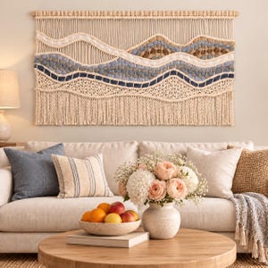 Peut inclure: Une grande tenture murale en macramé rectangulaire présentant un motif de paysage avec des couches ondulées de cordes nouées crème, bleu clair, bleu foncé et marron. L'œuvre est suspendue à une tige en bois et terminée par une longue frange.