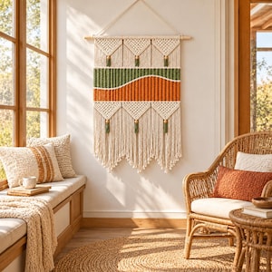Peut inclure: Une tenture murale en macramé composée de cordes couleur crème, d'une bande horizontale vert olive et d'une section ondulée orange brûlé. L'ouvrage est suspendu à une tige en bois et agrémenté de perles en bois et de pompons verts.