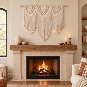 Puede incluir: Un tapiz de macramé grande en color blanco roto con intrincados patrones de nudos y largos flecos fluidos, colgado de una vara de madera. La pieza se exhibe sobre una repisa de chimenea de piedra con fuego y objetos decorativos.