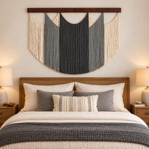 May include: A large macrame wall hanging featuring a dark wooden horizontal bar at the top. The piece consists of long, vertical cotton cords arranged in a layered, scalloped pattern, transitioning in colour from cream to light grey to dark charcoal.