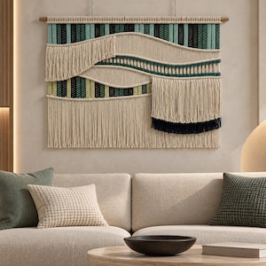 Puede incluir: Un gran tapiz de macramé rectangular colgado de un listón de madera. La pieza presenta un diseño en capas con cordón color crema, intercalado con rayas verticales en verde azulado, verde oscuro y verde lima, acabado con largos flecos.