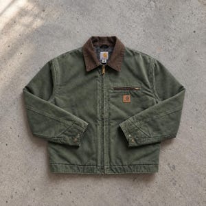 May include: An olive green jacket with a brown corduroy collar and a full-length front zip. The jacket features a horizontal zipped chest pocket, a square Carhartt brand patch, long sleeves with buttoned cuffs, and a size M label inside the collar.
