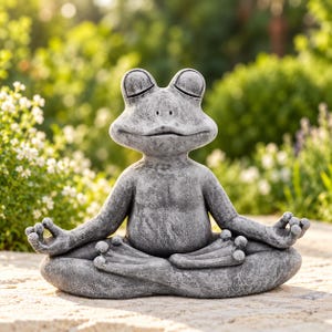 Peut inclure: Une figurine de grenouille à la texture de pierre grise dans une pose de méditation assise. La grenouille a les yeux fermés, une large bouche et des pattes palmées en position jambes croisées, les mains posées sur les genoux en mudra.