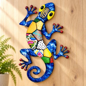 Könnte beinhalten: Eine Wandskulptur in Form eines Geckos mit einem leuchtend blauen Körper in Marmoroptik und hellgelben Augen. Der Rumpf ist mit einem Patchwork aus bunten geometrischen und floralen Mustern in Orange-, Grün- und Weißtönen verziert.