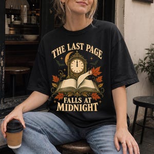 Pode incluir: Uma t-shirt preta com uma estampa gráfica de um livro aberto com um relógio por cima, rodeado por folhas de outono, morcegos e uma lua crescente. O texto diz THE LAST PAGE FALLS AT MIDNIGHT numa fonte serifada clara e em destaque.