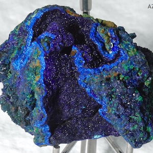May include: A raw azurite mineral specimen featuring deep royal blue and vibrant green crystalline clusters with a textured, jagged surface. The specimen is labeled with the text AZR-001 in the upper right corner and is mounted on a metal stand.