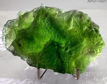 May include: A vibrant, translucent green mineral specimen with a jagged, crystalline texture, displayed on a small metal stand. The upper right corner features the text APO-001. The stone exhibits varying shades of bright lime and emerald green.
