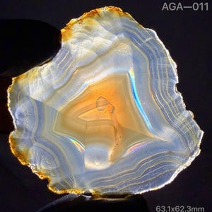 May include: A translucent agate slice with concentric bands of white, blue, and amber, featuring a glowing golden center. The specimen is labeled AGA-011 and measures 63.1x62.3mm, displaying natural mineral formations and light refraction.