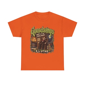 May include: An orange short-sleeve t-shirt featuring a graphic print of the Goosebumps Night of the Living Dummy book cover. The illustration shows a ventriloquist dummy in a chair, a lamp, books, and blocks spelling SLAPPY, with R.L. STINE text.
