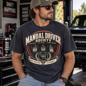 May include: A charcoal gray t-shirt featuring a graphic of three car pedals and a manual gear shift pattern. The text reads Manual Driver Society, Three Pedals, Real Drivers, A Higher Standard, More Than A Drive, and It's A Mindset.