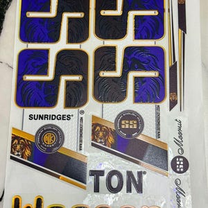 Könnte beinhalten: Ein Bogen mit Cricket-Schläger-Aufklebern, die das SS-Logo in Lila und Schwarz mit goldenen Umrandungen zeigen, sowie die Schriftzüge SUNRIDGES, TON und klaasen und kreisförmige Embleme mit Löwenkopf und dem Text PREMIUM ENGLISH WILLOW.
