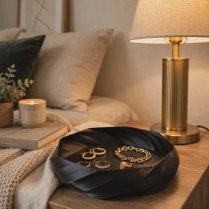 May include: A black circular tray with a twisted, rippled edge design holding a collection of gold jewelry, including two small hoop earrings, a thick chain link bracelet, a thin band ring, a beaded ring, and a signet ring on its flat surface.